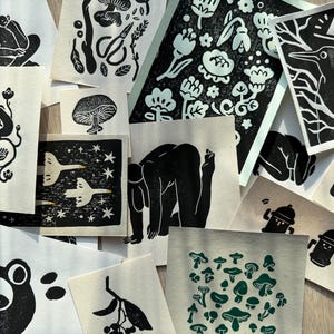 Może przedstawiać: Kolekcja czarno-białych linorytów przedstawiających różne wzory, w tym grzyby, kwiaty, ptaki i abstrakcyjne kształty. Drzeworyty są prezentowane na jasnobrązowej powierzchni.