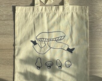 Puede incluir: Una bolsa de tela beige con una ilustración en blanco y negro de una persona sosteniendo un hongo. La persona lleva un sombrero de hongo y está rodeada de cuatro hongos.