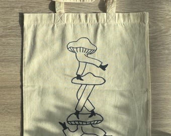 Puede incluir: Una bolsa de tela beige con asas negras y una ilustración en blanco y negro de una persona hongo de pie sobre otra persona hongo.