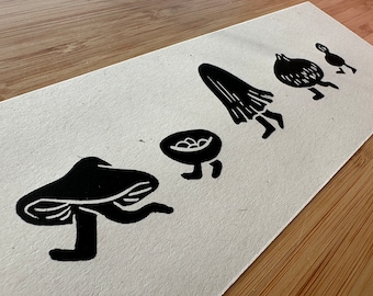 May include: Black and white linocut print featuring whimsical illustrations of a mushroom, a bowl with eggs, a ghost, an onion, and a small bird, all with legs, as if running. The print is on textured white paper, set against a wooden surface.
