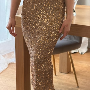 May include: A gold sequin skirt. The skirt is knee-length and covered in small, round, gold sequins. The skirt appears to be made of a knit material. The skirt is worn with a gray top and dark burgundy boots.