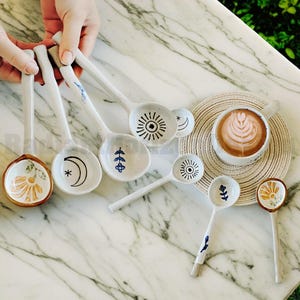 May include: A set of nine white ceramic spoons with various hand-painted designs, including a sun, moon, flowers, and leaves. The spoons are arranged on a white marble surface with a cup of coffee and a woven mat.