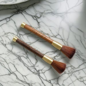 May include: Two wooden makeup brushes with gold accents and natural bristles. The brushes have engraved text on the handles, "The Art of the Brush" and "Coffee with a Brush".