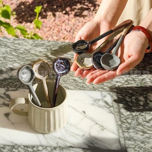 May include: A set of six ceramic spoons in various colours, including black, grey, and white, with a speckled glaze. The spoons are displayed in a white ceramic mug with a textured surface.