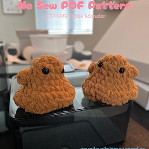 May include: Two handmade, plush, orange-brown SCP-999 Tickle Monster toys with black button eyes. The toys are sitting on a black surface. The text "No Sew PDF Pattern" and "SCP-999 Tickle Monster" is visible in the background.