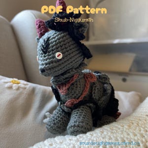 Può includere: Una creatura grigia all'uncinetto con dettagli neri e rosa, con un occhio a bottone e un colletto decorativo rosa e marrone. Il testo "PDF Pattern Shub-Niggurath" è visualizzato nell'immagine. La creatura è su una superficie bianca.