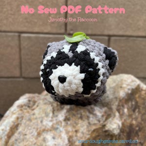 May include: A crochet raccoon plush toy with grey, black, and white yarn, featuring black plastic eyes and a small green leaf on top. The text reads No Sew PDF Pattern Jimothy the Raccoon and the website address sourdoughgames.carrd.co.