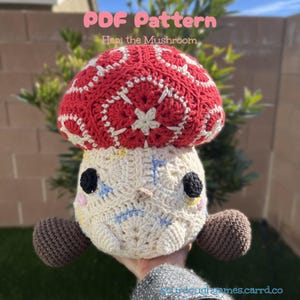 Può includere: Peluche a forma di fungo all'uncinetto con un cappello a fantasia rosso e bianco e un corpo color crema. Il fungo ha occhi neri, un naso piccolo e gemme laterali marroni. Il testo "PDF Pattern Hapi the Mushroom" è visibile.
