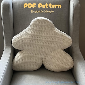 May include: A cream-colored, crocheted pillow in the shape of a meeple, resting on a gray armchair. The pillow has a unique, rounded shape with a textured surface. Text at the top reads "PDF Pattern Huggable Meeple".