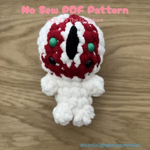 May include: A crocheted plush toy featuring a white body and a textured head with red and white yarn, a central black vertical eye, two small green circular accents, and two black bead eyes. The text reads No Sew PDF Pattern SCP-173 The Sculpture.