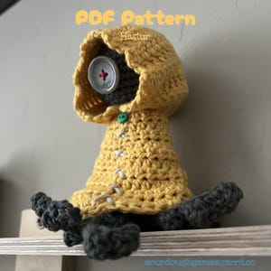 Pode incluir: Boneca de crochê feita à mão em uma capa com capuz amarelo com um grande olho de botão e membros cinzentos. A boneca tem detalhes de costura brancos e um fecho de botão verde. O texto "PDF Pattern Hastur" é visível no topo.