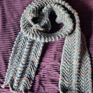 May include: A hand-knitted scarf in shades of blue, green, and pink. The scarf features a textured chevron pattern and is displayed on a purple fabric surface. The scarf is a cozy accessory.