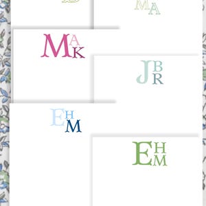 May include: A collection of personalized stationery cards with various monogram designs. The cards feature initials in different colors, including pink, blue, and green, set against a white background. The background has a floral pattern.