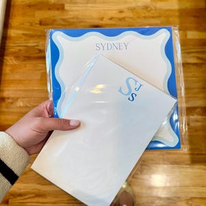 May include: Two personalized stationery sets. One set features the name "SYDNEY" in blue on a white background with a blue scalloped border. The other set has the initials "SJ" in blue. Both sets are packaged in clear plastic.