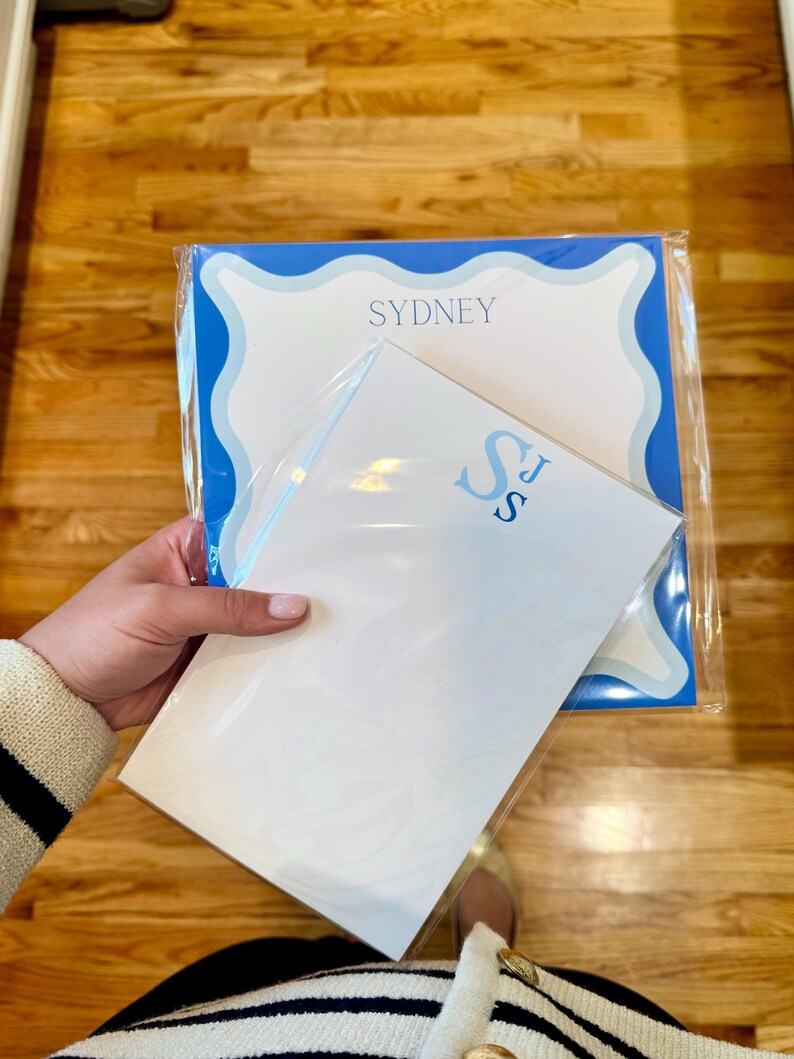 May include: Two personalized stationery sets. One set features the name "SYDNEY" in blue on a white background with a blue scalloped border. The other set has the initials "SJ" in blue. Both sets are packaged in clear plastic.