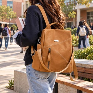Pode incluir: Uma mochila conversível castanho-clara com alça superior e alças de ombro ajustáveis. A bolsa possui um bolso frontal vertical com zíper, uma pequena placa metálica retangular e detalhes de costura diagonal. Acabamento liso.