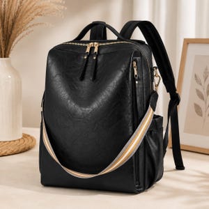 Puede incluir: Una mochila negra con acabado texturizado, cremalleras doradas y una correa de hombro extraíble con rayas blancas, grises y bronceadas. Incluye un asa superior, correas de hombro ajustables y bolsillos laterales para almacenamiento.