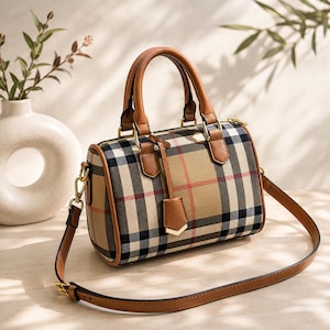 May include: A barrel-shaped handbag featuring a tan, black, and red plaid pattern. The bag has brown leather handles, a matching adjustable shoulder strap, gold-tone metal hardware, and a small leather tag hanging from the front handle base.