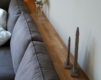 May include: A long, narrow, rectangular wooden console table shelf positioned directly behind a grey fabric sofa. The light brown wood surface features a visible grain pattern and holds two silver-toned decorative goblets and two tall, thin, pointed wooden sculptures.