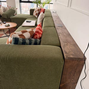 Może przedstawiać: Duża, oliwkowozielona sofa narożna z sztruksu z ciemnym drewnianym podłokietnikiem. Sofa jest ozdobiona różnymi poduszkami w odcieniach bieli, brązu i czerwieni. W tle widoczny jest drewniany stolik i okno.