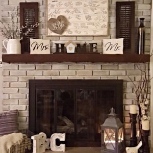 May include: A fireplace mantel display featuring a white guest book sign with a heart monogram, dark brown shutters, a white pitcher with branches, and wooden blocks spelling HOME with a house icon. Includes Mrs. and Mr. signs and a lantern.