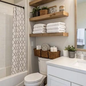 Puede incluir: Interior de baño con paredes blancas, mueble de lavabo blanco y un inodoro. Estantes de madera con toallas, plantas y objetos decorativos. Cestas de mimbre colocadas encima del inodoro. Se ve una cortina de ducha con estampado.