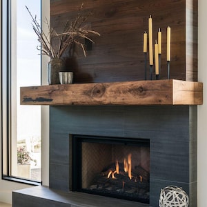 Op de afbeelding: Een moderne open haard met een donkergrijze betegelde omlijsting en een rustieke houten mantel. De open haard heeft een brandend vuur, een decoratieve metalen bol en een vaas met takken. De muur achter de mantel is gemaakt van horizontale houten planken.