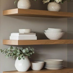 May include: Three wooden shelves with white ceramic vases filled with white flowers and greenery. The shelves are decorated with white ceramic bowls and plates.