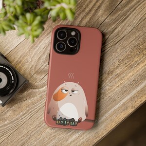 May include: A terracotta-colored phone case featuring a cartoon character with a sleepy expression. The character is a mix of beige, white, and orange, with the text "DAY BY DAY" at the bottom. Steam lines above the character add to the sleepy theme.