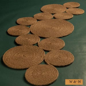 Puede incluir: Un conjunto de posavasos y un camino de mesa de cuerda marrón, dispuestos sobre una superficie verde oscuro. Los posavasos y el camino de mesa están hechos de cuerda enrollada, creando un diseño circular texturizado. La esquina inferior derecha tiene la inscripción "W & N".