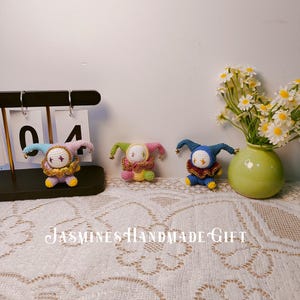 May include: Three handmade crochet jester dolls in various colors: pink, green, and blue. Each doll has a round body, a ruffled collar, and a pointed hat. The dolls are displayed on a lace tablecloth with the text "Jasmines Handmade Gift".