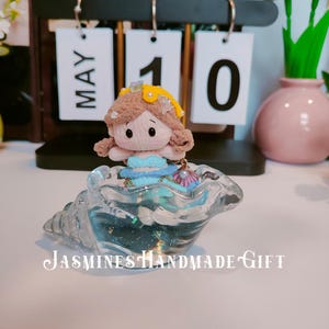May include: A handmade doll with a yellow crown and blue dress sits inside a clear, seashell-shaped container. The container has a glittery, blue interior. The text "Jasmines Handmade Gift" is visible. A calendar shows "MAY 10".