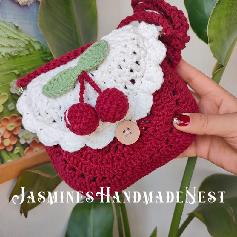 May include: A crocheted cherry-themed handbag with a white and red design. The bag has a white crocheted flower with a red cherry in the center. The bag has a red crocheted strap.