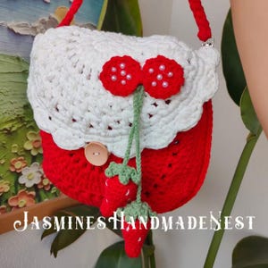 May include: A crocheted strawberry-shaped handbag with a white crocheted top and a green stem. The bag has a button closure and a long strap. The text "JASMINES HANDMADENEST" is visible at the bottom of the image.