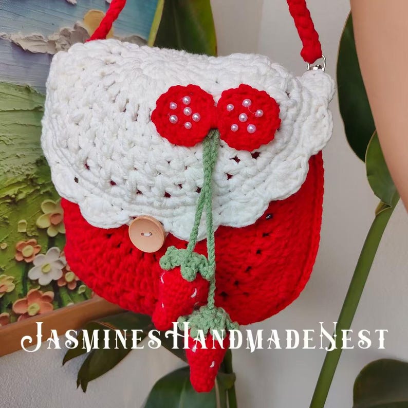 May include: A crocheted strawberry-shaped handbag with a white crocheted top and a green stem. The bag has a button closure and a long strap. The text "JASMINES HANDMADENEST" is visible at the bottom of the image.