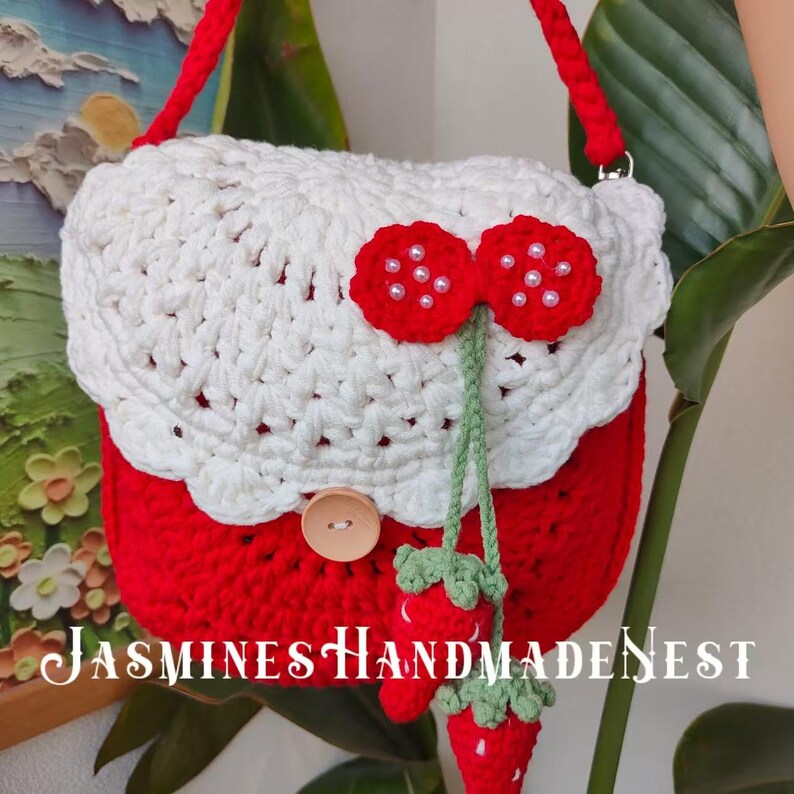 May include: A red and white crocheted handbag with a strawberry design. The bag has a white crocheted flap with a red strawberry design and a wooden button. The strap is red and white.