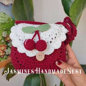 May include: A small red and white crocheted handbag with a cherry design. The bag has a white crocheted trim and a green crocheted bow. The bag has a brown button.