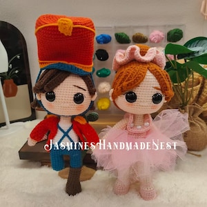 May include: A crocheted toy soldier wearing a red and blue hat and a blue and red uniform. Next to the soldier is a crocheted ballerina wearing a pink tutu and a white headband. Both toys have large black eyes and brown hair.