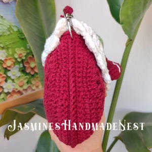 May include: A crocheted burgundy purse with a white crocheted trim and a silver clasp. The purse is held in a hand.