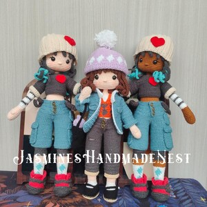 May include: Three handmade crochet dolls. Each doll wears a hat with a heart, a gray top, blue pants, and red and gray shoes. The dolls have different skin tones and hair colors. The text "JASMINE'S HANDMADE NEST" is at the bottom.