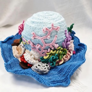 May include: A light blue crocheted hat with a wide, wavy brim. The crown is decorated with various three-dimensional crocheted marine elements, including pink coral, green and purple seaweed, white shells, and a single small white pearl bead.