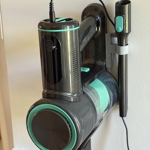 以下が含まれることがあります： グレーと黒の色合いで、ティールでアクセントを効かせたコードレス掃除機。掃除機は壁掛けブラケットに取り付けられており、充電ケーブルが含まれています。デザインは円筒形のダストビンと取り外し可能なノズルを備えています。