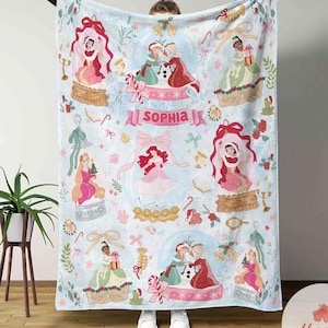 Puede incluir: Una manta azul claro con ilustraciones de princesas dentro de globos de nieve, con el nombre "Sophia" en una pancarta rosa. El dise&ntilde;o incluye bastones de caramelo, lazos y elementos decorativos, creando un tema caprichoso y festivo.