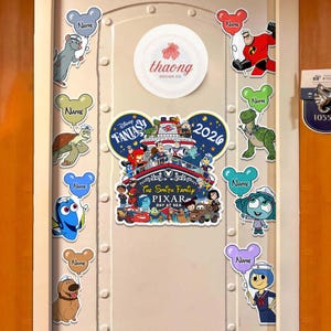 May include: A white door decorated with Disney-themed stickers. The stickers include characters like Dory, Remy, and Mr. Incredible, each holding a balloon with the word "Name." A central sticker reads "Disney Fantasy Pixar Day at Sea 2026."