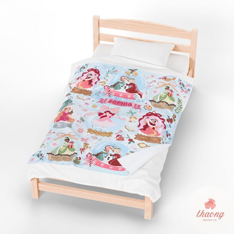 Puede incluir: Una manta azul claro con un patr&oacute;n repetido de bolas de nieve, cada una con una ilustraci&oacute;n diferente de una pareja o persona. La manta tiene el nombre "Sophia JK" impreso en rosa. La manta est&aacute; sobre una estructura de cama de madera.