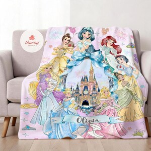 May include: A soft fleece blanket featuring a colorful illustration of multiple animated princesses surrounding a central castle graphic. The design includes a large blue ribbon, fireworks, and a banner with the name Olivia printed in black script.