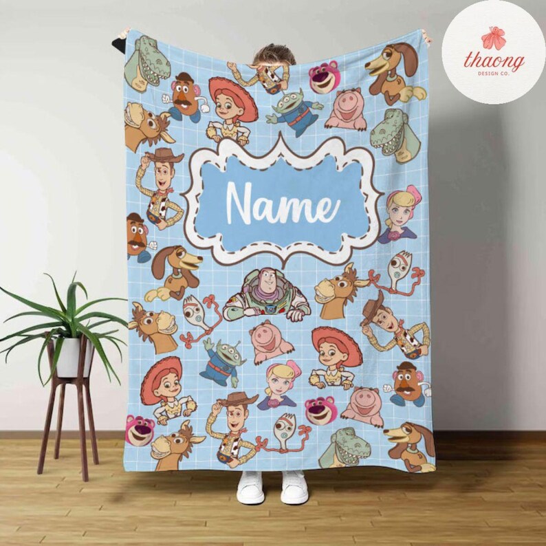May include: A blue fleece blanket with a grid pattern and a variety of Toy Story characters, including Woody, Buzz Lightyear, Jessie, and Mr. Potato Head. The blanket has a blue frame with the word "Name" in white text.
