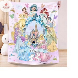 May include: A light purple fleece blanket featuring a central illustration of a castle surrounded by various animated princesses in colorful gowns. The design includes fireworks, stars, and balloons, with a blue ribbon banner displaying the name Olivia.