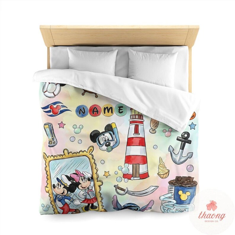 Puede incluir: Un edred&oacute;n colorido con un dise&ntilde;o caprichoso con Mickey y Minnie Mouse, un faro, un ancla y otras ilustraciones de tem&aacute;tica n&aacute;utica y Disney. Tambi&eacute;n se muestra la palabra "NAME". El edred&oacute;n est&aacute; sobre una cama con almohadas blancas.