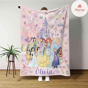 May include: A pink fleece blanket with a colorful illustration of Disney princesses in front of a castle. The blanket has a watercolor effect and features a pink and purple color scheme. The text "Olivia" is printed in purple at the bottom of the blanket.